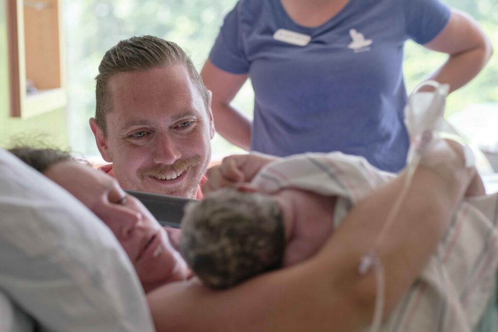 dad looks at newborn and mom after birth in hospital with doula behind them