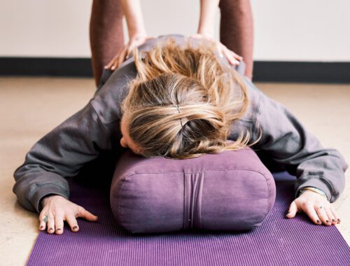 A person resting on a bolster, receiving an adjustment from a Yoga teacher.