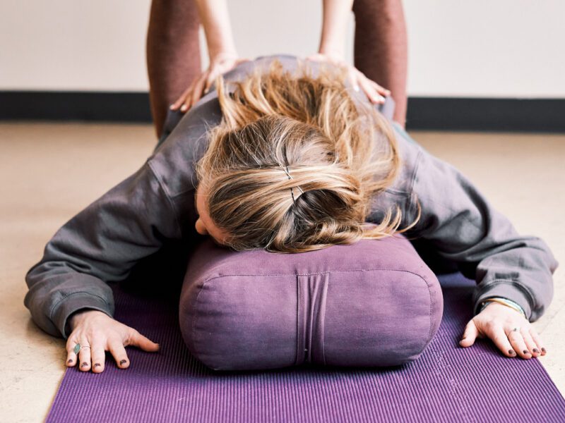 A person resting on a bolster, receiving an adjustment from a Yoga teacher.