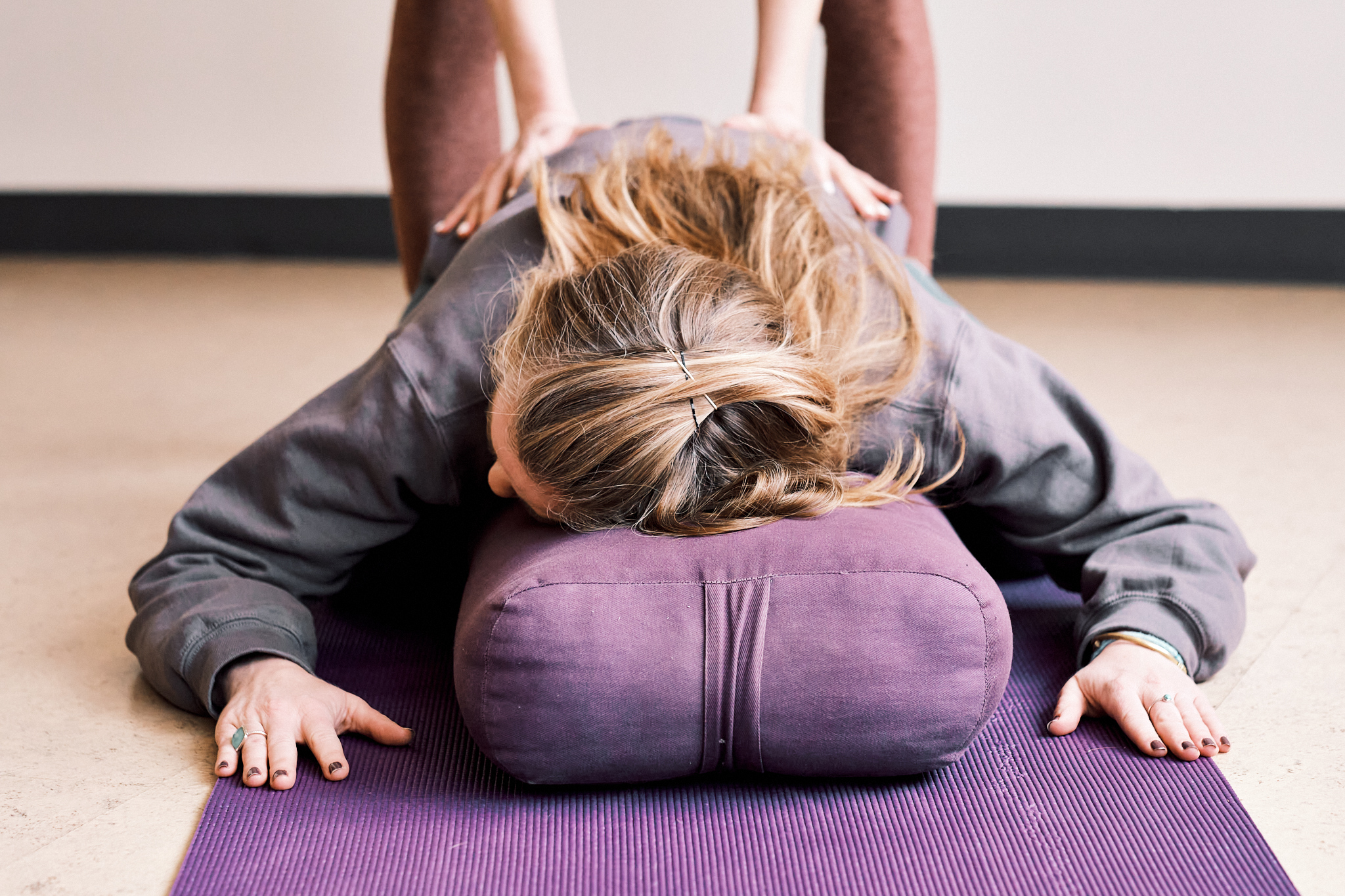 A person resting on a bolster, receiving an adjustment from a Yoga teacher.