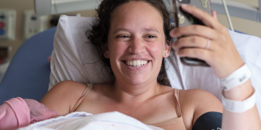 new mom in hospital bed with newborn video chatting with a friend